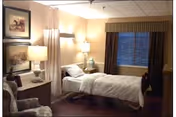 A nursing facility bedroom with a single bed, bedside tables and lamps, an armchair, framed art, and a window with curtains.
