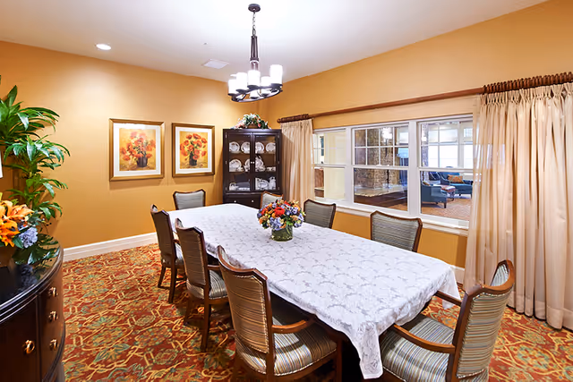A dining room with a long rectangular table covered with a white lace tablecloth and surrounded by eight wooden chairs with striped cushions. A floral centerpiece is placed in the middle of the table. The room has warm beige walls, a patterned carpet, two framed floral paintings on the wall, a dark wooden cabinet displaying plates, and a window with beige curtains looking into another room with seating.