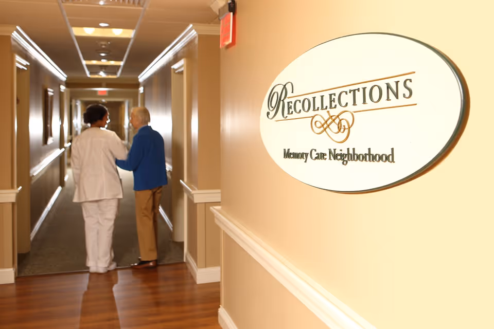 A hallway in a senior living facility with beige walls and wooden flooring. A sign on the right wall reads 'Recollections Memory Care Neighborhood.' Two people, one in white medical attire and the other an elderly person in a blue sweater, are walking down the hallway together.