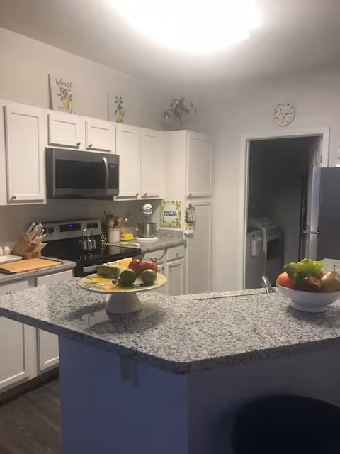 Modern kitchen with white cabinets, granite countertops, stainless steel microwave and stove, a bowl of fruit on the counter, and kitchen utensils. There is a doorway leading to another room with a refrigerator visible.