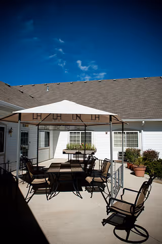 Outdoor patio area at Countryside Meadows with a white canopy covering a table and several chairs arranged around it. The patio is surrounded by white buildings with windows and potted plants, under a clear blue sky.