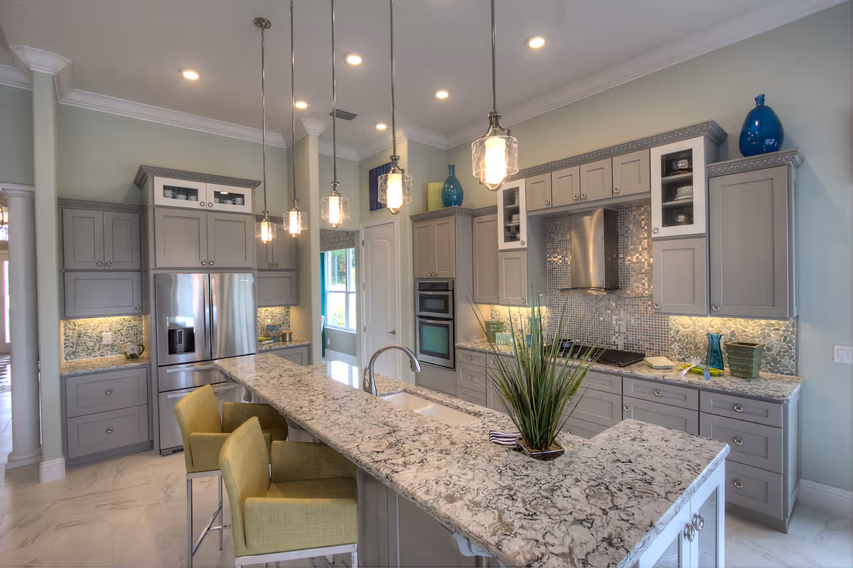 Modern kitchen with gray cabinets, stainless steel refrigerator, built-in oven, and a large granite countertop island with a sink and green upholstered chairs. Pendant lights hang above the island, and decorative vases are placed on top of the cabinets.