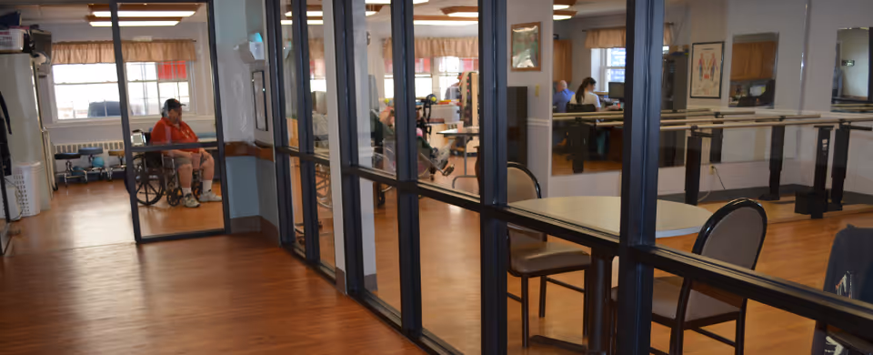 Interior view of a rehabilitation facility room with wooden floors and glass partitions. Several people are seated, some in wheelchairs, engaging in activities or resting. There are tables and chairs arranged in the room, and exercise equipment is visible in the background.