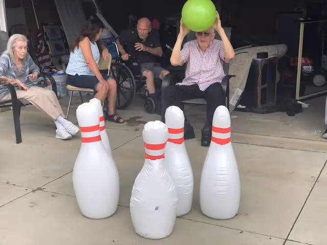 A group of elderly people sitting outside on chairs and in a wheelchair, playing a game with large inflatable bowling pins and a green ball. One man is holding the green ball above his head, preparing to roll it towards the pins.