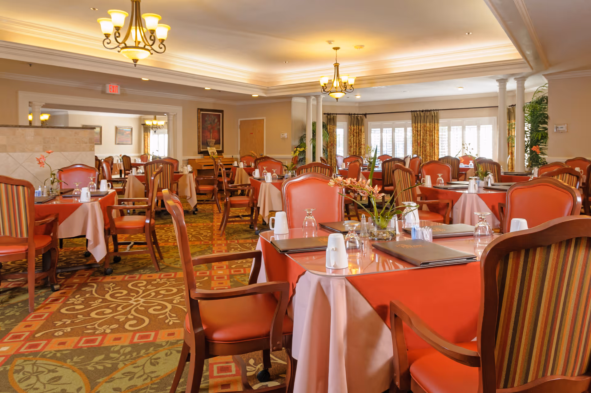 A spacious dining room with multiple tables covered in red and beige tablecloths, set with glasses, salt and pepper shakers, and menus. The room features wooden chairs with red and striped upholstery, chandeliers hanging from the ceiling, patterned carpet, and large windows with curtains allowing natural light to enter.