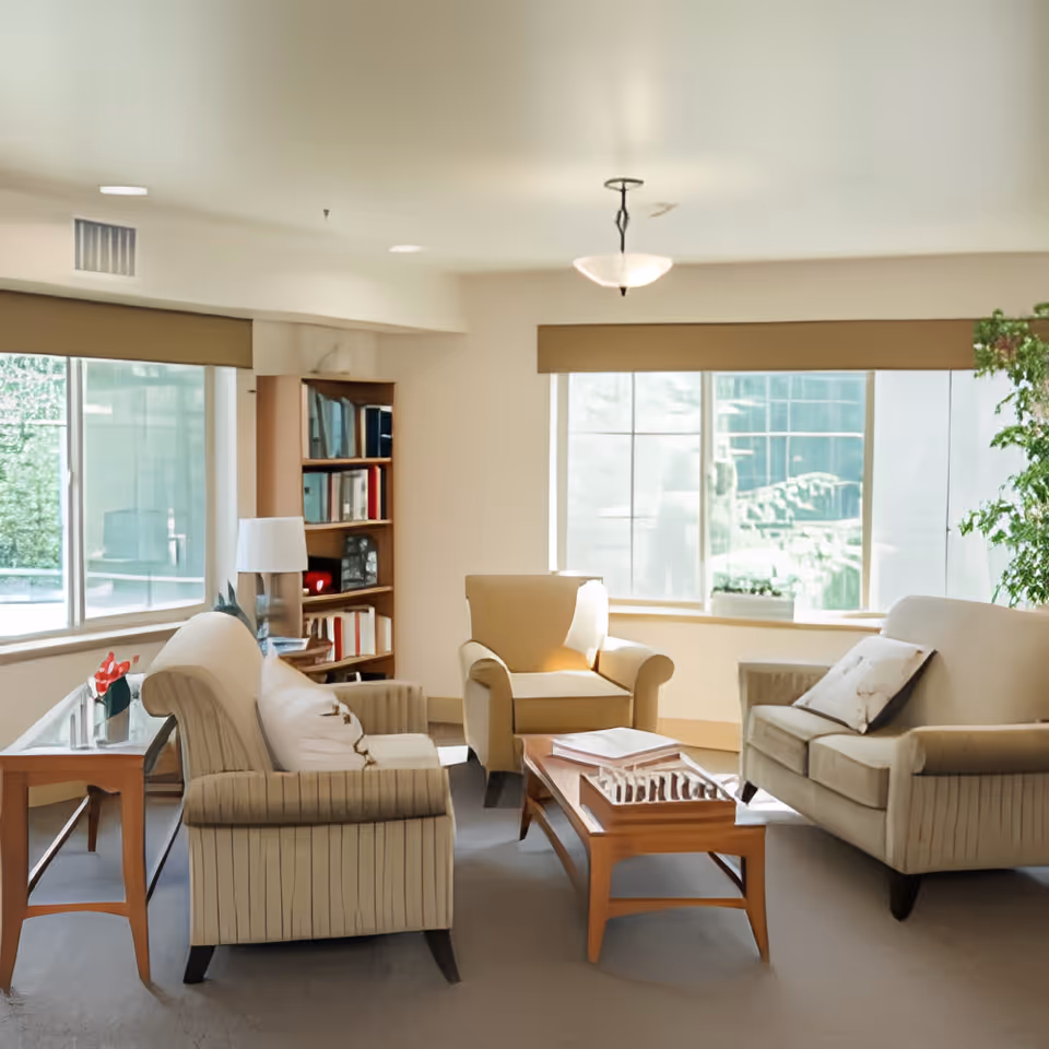 A bright and cozy living room with large windows letting in natural light. The room features a beige armchair, a matching loveseat with a pillow, a striped sofa, a wooden coffee table with books and a decorative item, a side table with a lamp, and a bookshelf filled with books. There is also a green plant near the window.