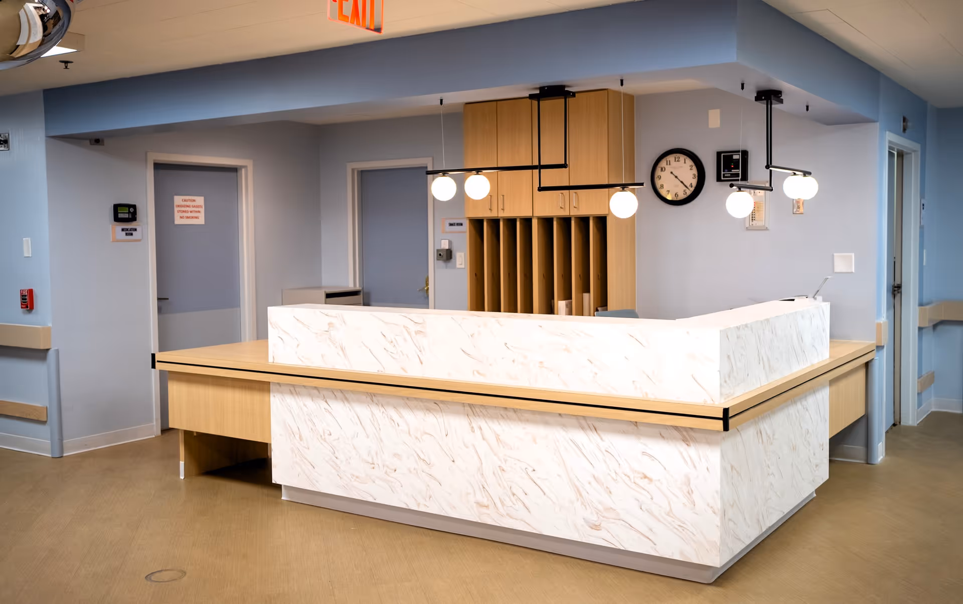 Reception desk area in a healthcare facility with a marble-patterned counter, wooden cabinetry, hanging globe lights, a wall clock, and several doors in the background.