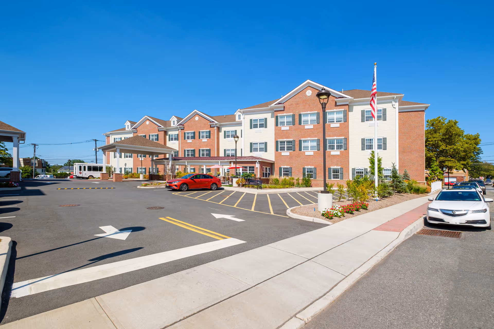 Exterior view of The Chelsea at Fair Lawn senior living facility showing a three-story brick and siding building with multiple windows, a covered entrance, a parking lot with cars, a flagpole with an American flag, and clear blue sky.