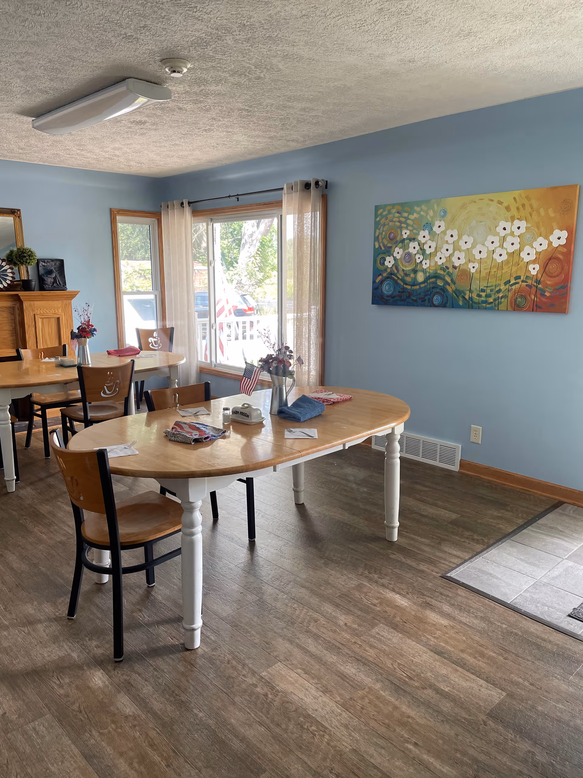Sunlit dining room with wooden tables and chairs, blue walls, a large window and colorful floral wall art.