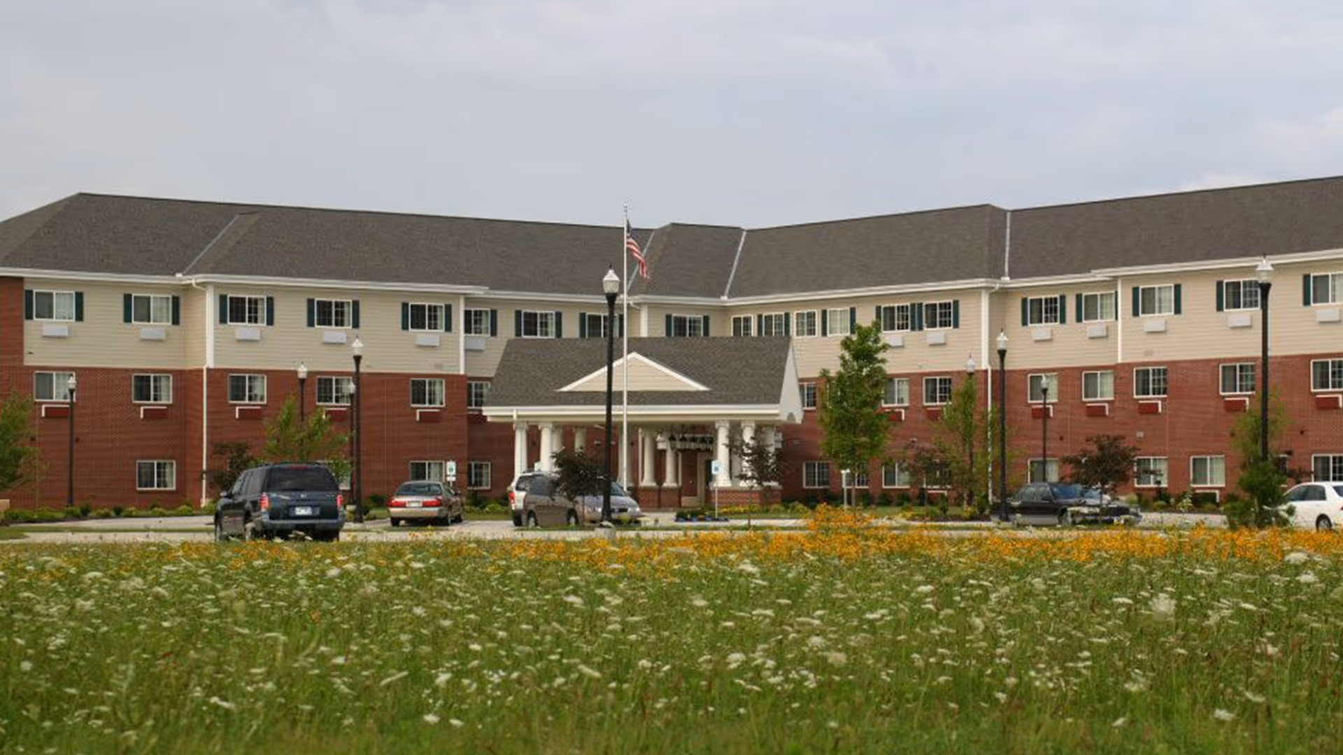 Front exterior of a multi-story brick and siding senior living building with a portico entrance, cars in the driveway, and a wildflower lawn in front.