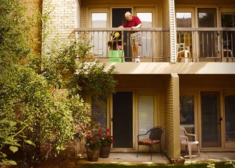 An elderly man watering plants on the balcony of a two-story residential building with beige siding. Below the balcony, there are two chairs and potted plants near the entrance doors. Green bushes and trees surround the building.