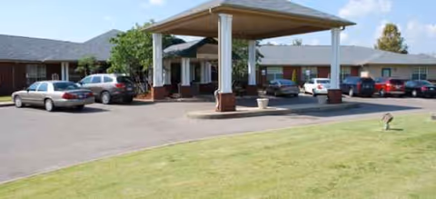 Exterior view of Shelby Gardens Place with a covered entrance, parked cars, and grassy foreground.