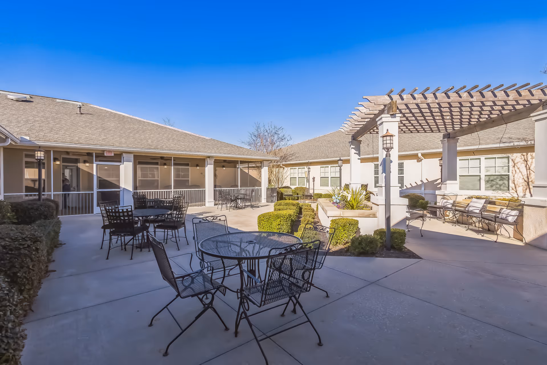 Outdoor courtyard area of a senior living facility with metal tables and chairs, a pergola with benches underneath, trimmed bushes, and a clear blue sky.