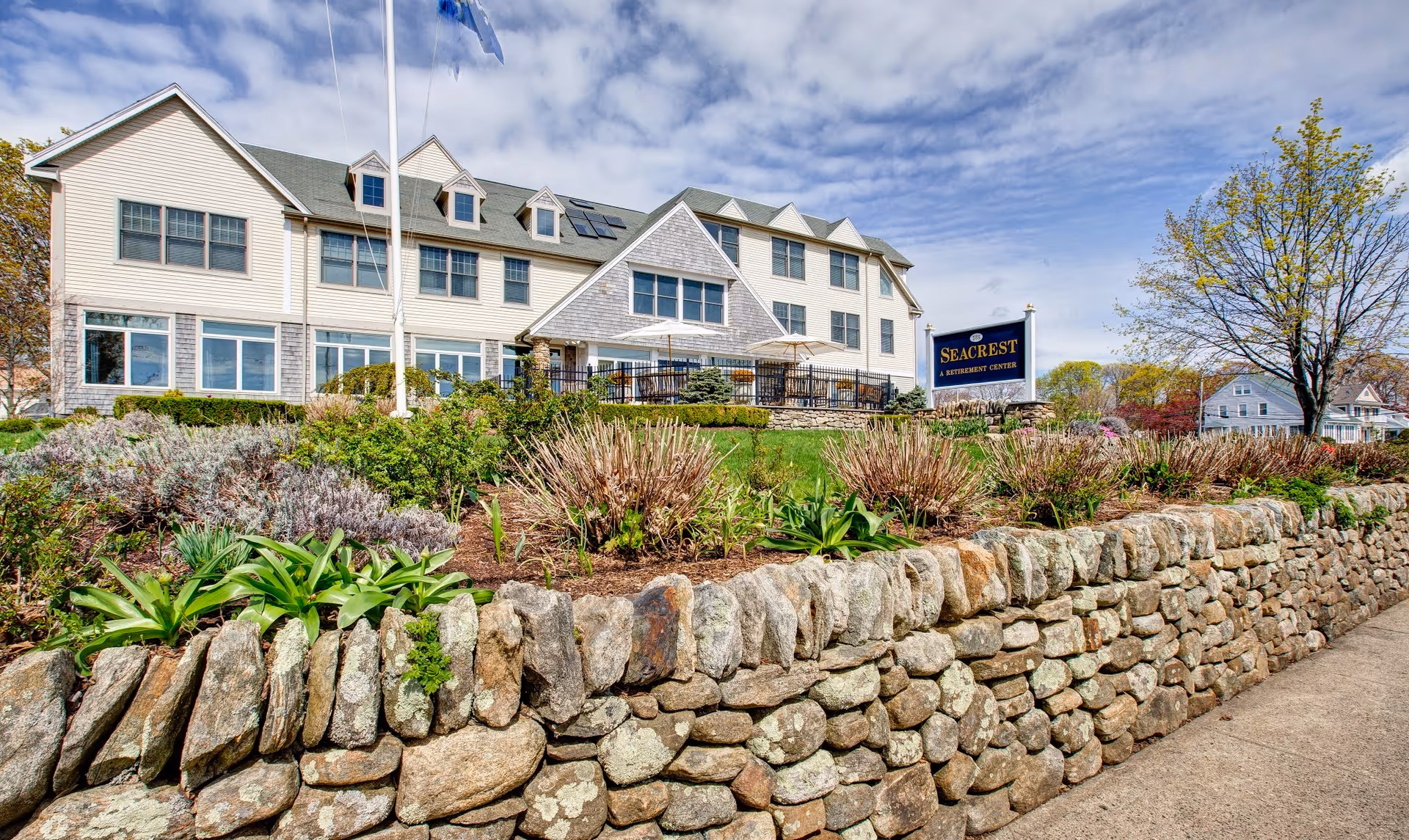 Stone retaining wall and landscaped garden leading up to the Seacrest Assisted Living & Memory Care building with its sign under a partly cloudy sky.