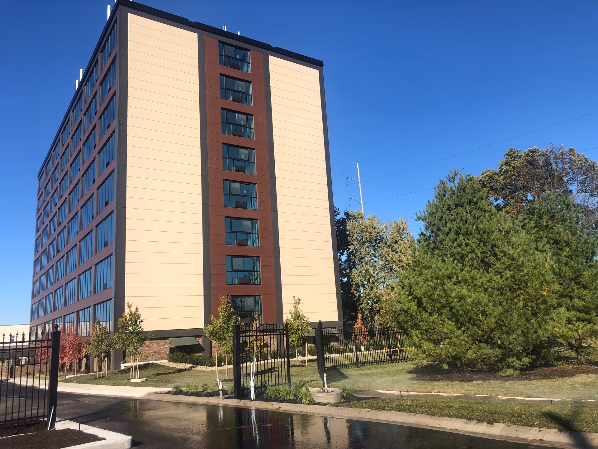 Exterior view of a multi-story senior living facility building with beige and brown panels, surrounded by a black metal fence, small trees, and green landscaping under a clear blue sky.