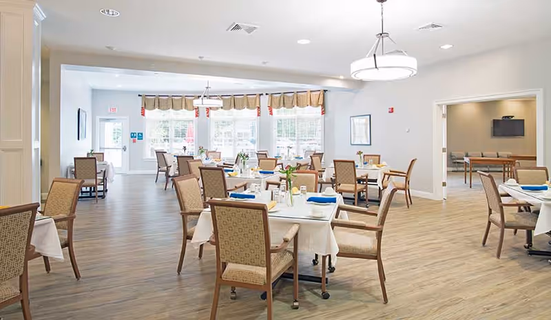 A bright and spacious dining room with multiple tables set with white tablecloths, blue napkins, and small flower vases. The room has large windows with valances letting in natural light, wooden flooring, and beige walls. There is an adjacent room visible through an open doorway with additional seating and a wall-mounted TV.