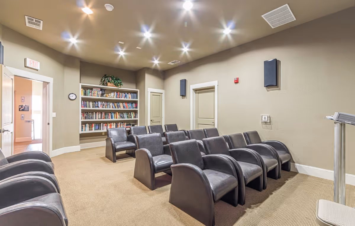 Small screening/library room with rows of black armchairs facing a podium, a built-in bookshelf, and recessed ceiling lights.