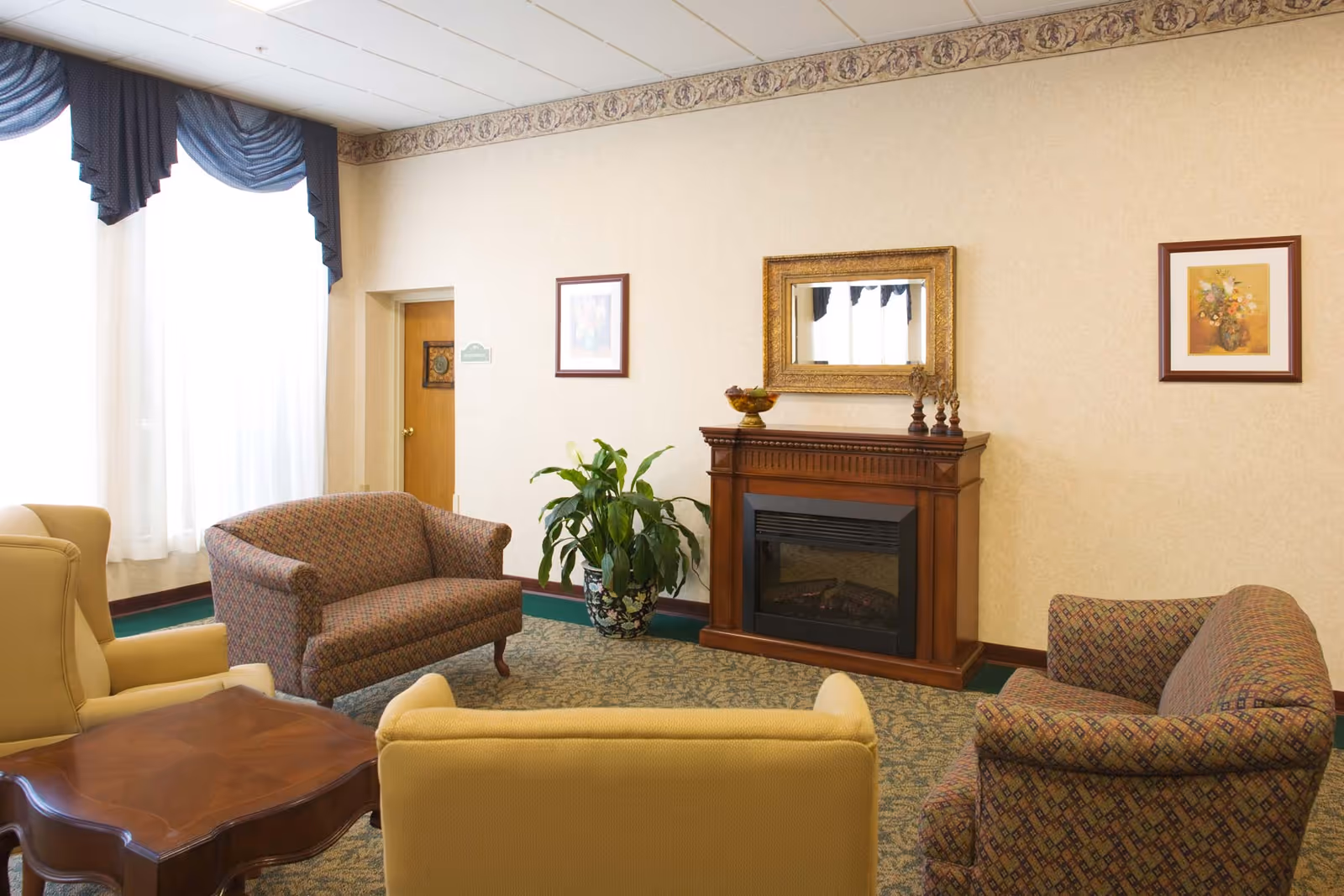 A cozy living room area with patterned sofas and yellow armchairs arranged around a wooden coffee table. The room features a wooden electric fireplace with a decorative mirror above it, framed floral artwork on the walls, a large potted plant, and a window with sheer white curtains and dark blue valances.