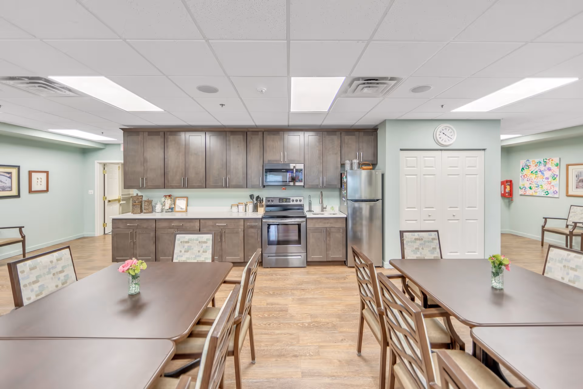A bright communal kitchen and dining area with wooden tables and chairs, a kitchen with dark wood cabinets, stainless steel appliances including a refrigerator and stove, and light green walls decorated with framed artwork and a clock above white double doors.