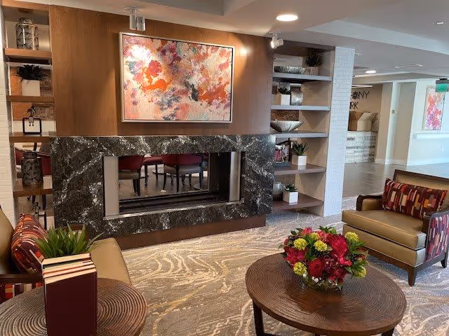 A cozy living room area with two armchairs featuring patterned cushions, a round wooden coffee table with a floral arrangement, and a modern double-sided fireplace with a black marble surround. The room has built-in shelves with decorative items and a colorful abstract painting above the fireplace. The background shows a hallway with additional artwork on the walls.