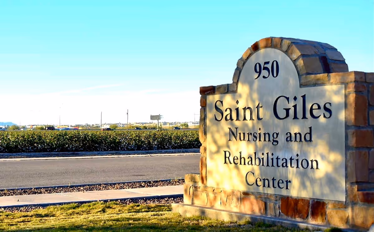 Stone sign for Saint Giles Nursing and Rehabilitation Center with the address number 950, situated near a road with greenery and a clear blue sky in the background.