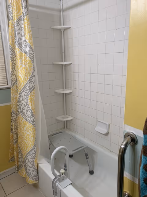 A bathroom with a white tiled bathtub and shower area. There is a yellow and gray patterned shower curtain partially drawn to the side. Inside the bathtub, there is a gray shower chair with adjustable legs and a white grab bar attached to the side of the tub. A vertical corner shelf with four tiers is mounted on the tiled wall. A metal grab bar is installed on the yellow-painted wall next to the tub.