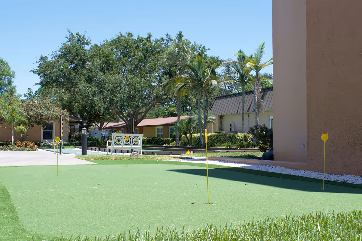 Outdoor putting green with several yellow golf hole flags, surrounded by grass and landscaping. In the background, there are palm trees, other greenery, a white bench, and buildings under a clear blue sky.