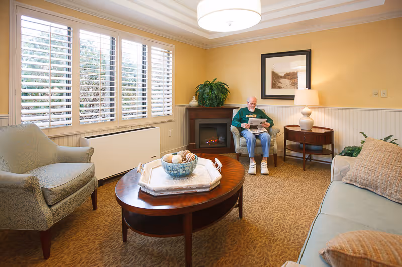 A cozy living room with beige walls and carpet, featuring a large window with white shutters, a wooden coffee table with decorative items, a fireplace with a green plant on top, a side table with a lamp, and an elderly man sitting in a chair reading a newspaper.
