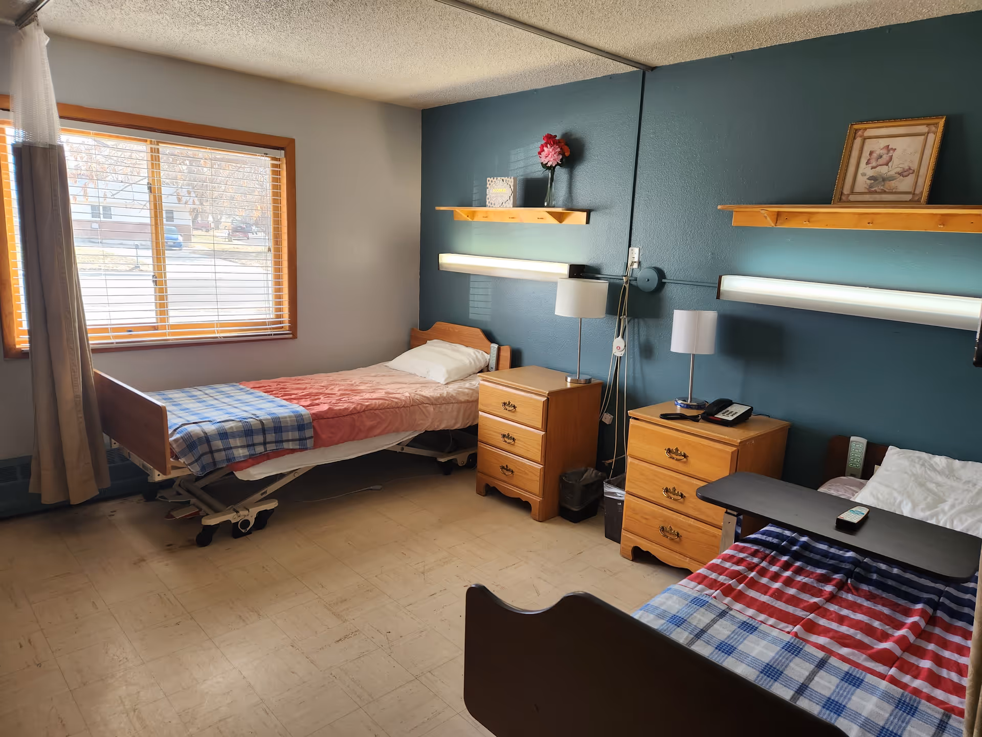 A shared room in a senior living facility with two single beds, each with a checkered blanket and a pillow. Between the beds are two wooden nightstands with lamps, a telephone, and a remote control. The walls are painted teal and beige, with wooden shelves holding decorative items. A large window with blinds lets in natural light.
