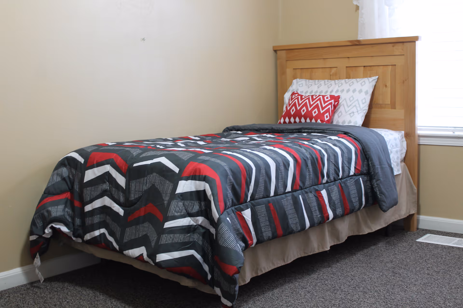 A single bed with a wooden headboard in a room with beige walls and carpeted floor. The bed is made with a comforter featuring a geometric pattern in red, black, white, and gray, and has two pillows, one with a matching geometric pattern and a smaller red decorative pillow. A window with white blinds is partially visible on the right side.