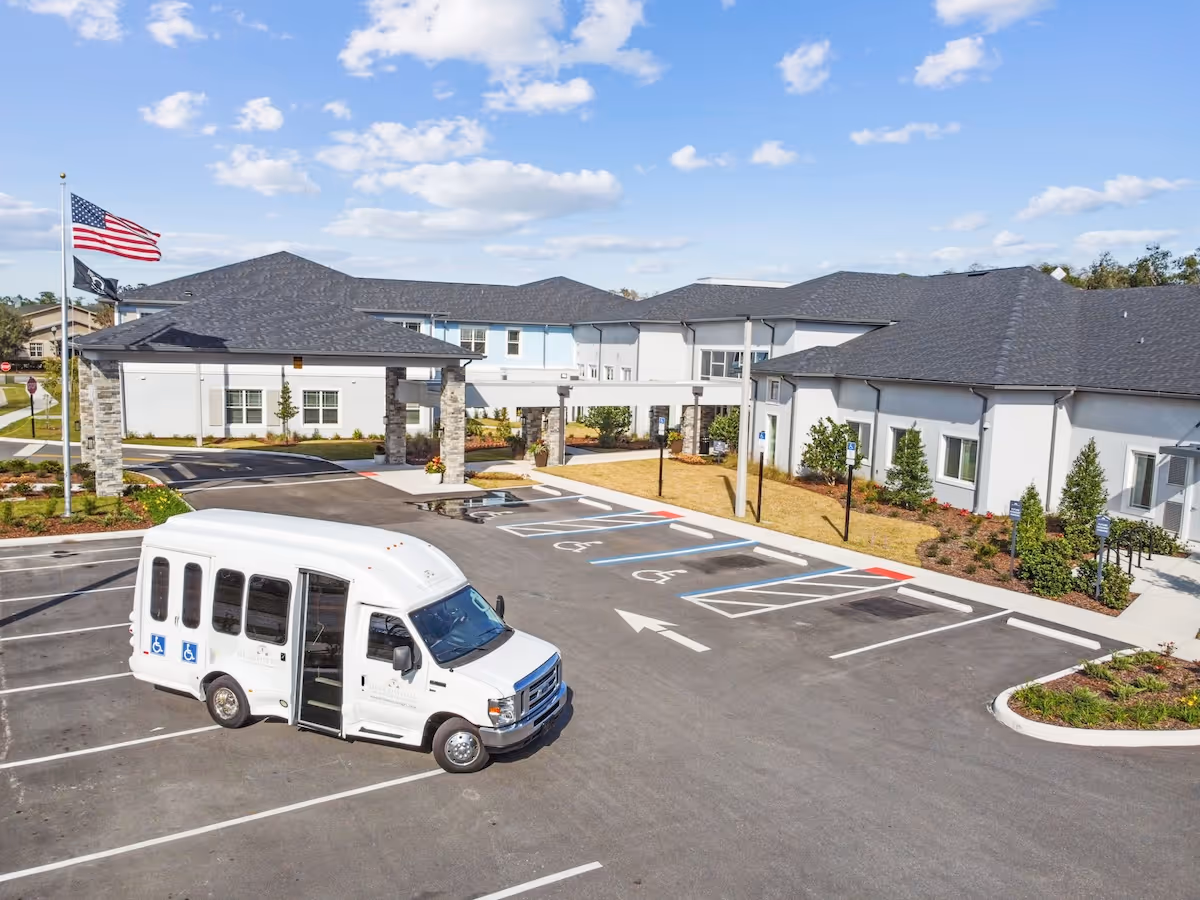 Exterior view of HearthStone at Nona Lakes senior living facility showing a white shuttle van parked in the parking lot with handicap parking spaces, a covered entrance supported by stone pillars, and an American flag flying on a flagpole under a partly cloudy sky.