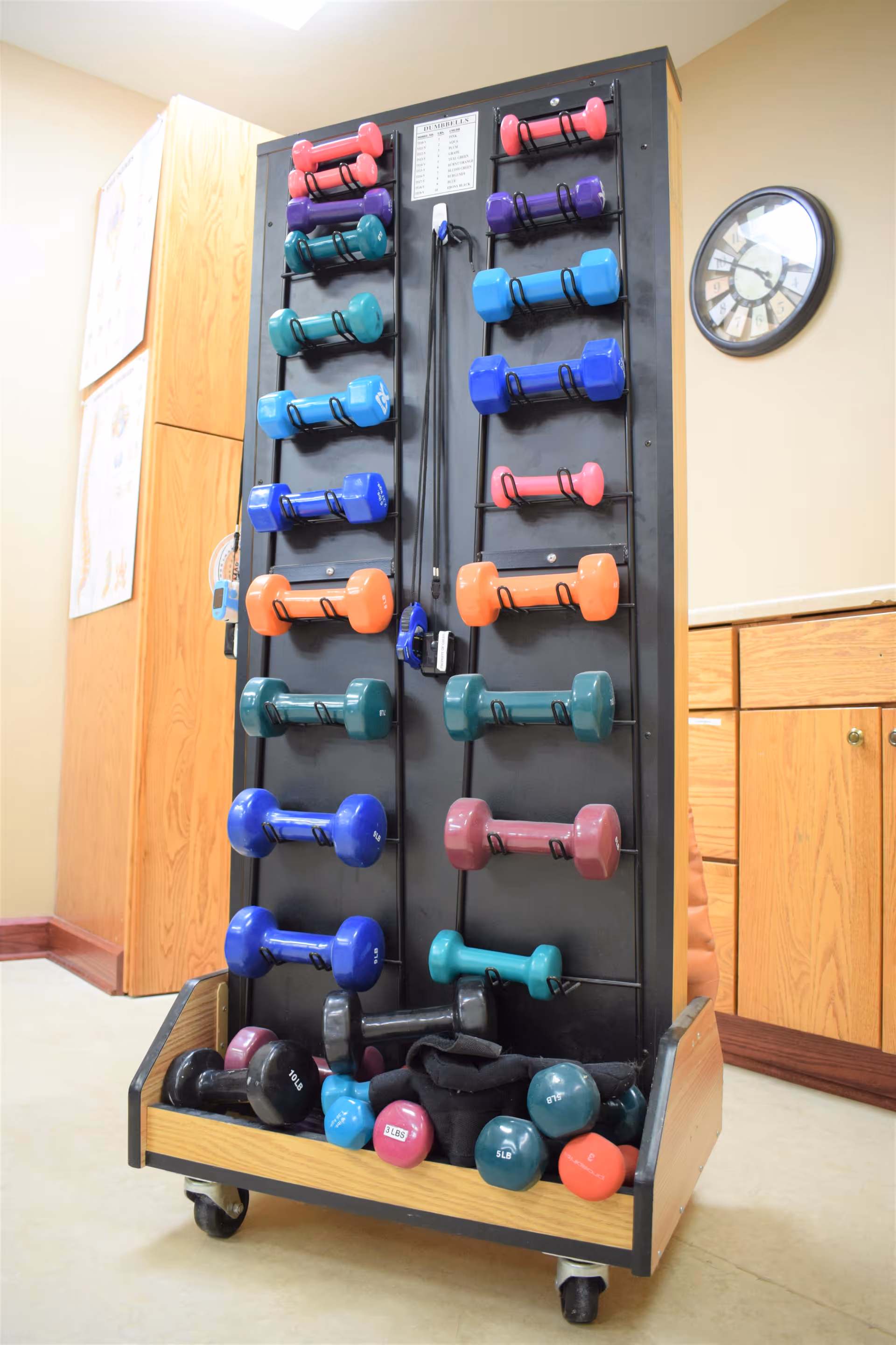 A black rack on wheels holding various colorful dumbbells of different weights, placed in a room with wooden cabinets and a wall clock.