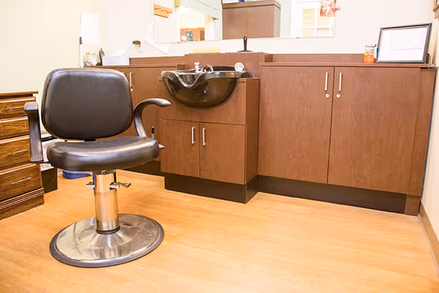 Interior of a salon area with a black salon chair in front of a wooden cabinet with a black sink. The floor is light wood, and there is a mirror and some framed items on the cabinet.