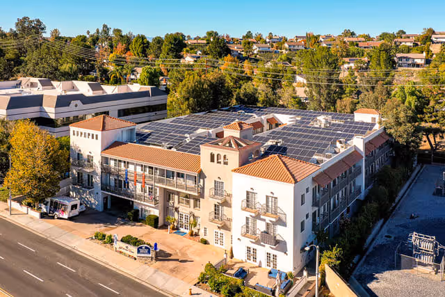 Aerial view of a multi-story senior living facility with solar panels on the roof, surrounded by trees and residential houses in the background. The building has a Mediterranean architectural style with balconies and a tiled roof.