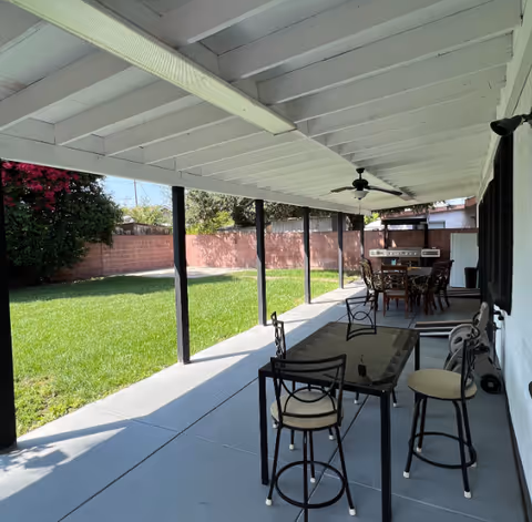 Covered backyard patio with multiple outdoor dining tables and chairs, ceiling fans, and a grassy lawn bordered by a brick wall.