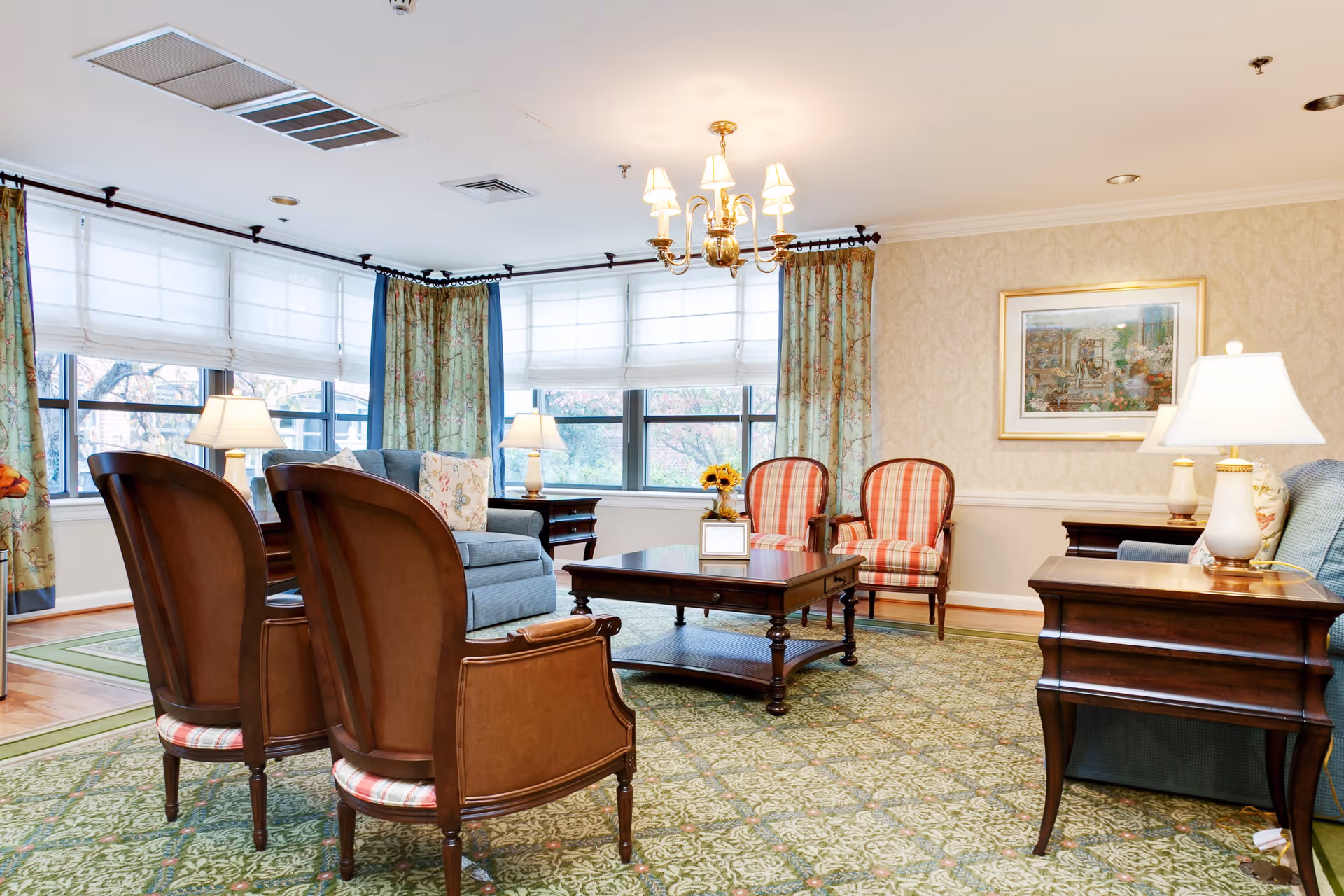 A bright and elegant living room with large windows covered by white blinds and floral curtains. The room features a blue sofa, two wooden armchairs with plaid cushions, two striped armchairs, a wooden coffee table, side tables with lamps, and a framed painting on the wall. The floor is covered with a patterned green carpet.