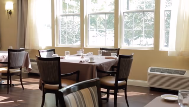 A bright dining room with large windows letting in natural light. Several tables are set with cups, glasses, and napkins, surrounded by wooden chairs with cushioned seats. The room has light-colored walls and dark wooden flooring.