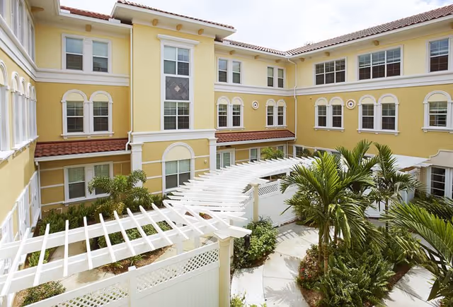 Sunlit yellow Mediterranean-style courtyard with a white pergola, palm plants, and surrounding multi-story building windows.