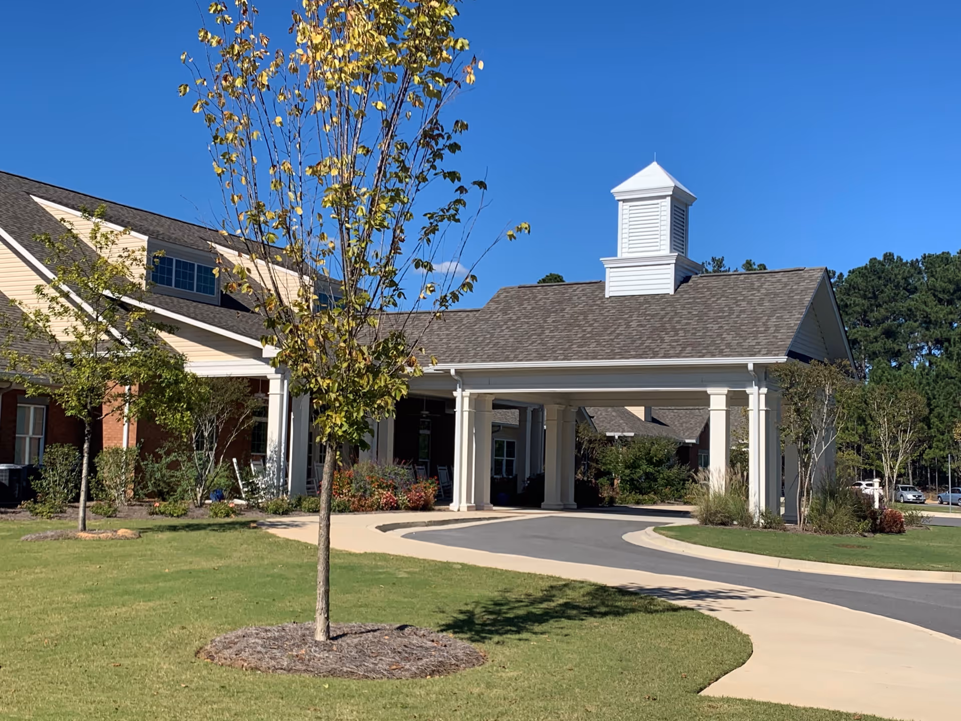 Exterior view of The Cottages of Milledgeville facility showing a covered entrance with white columns, a paved driveway, green lawn, young trees, and a clear blue sky.