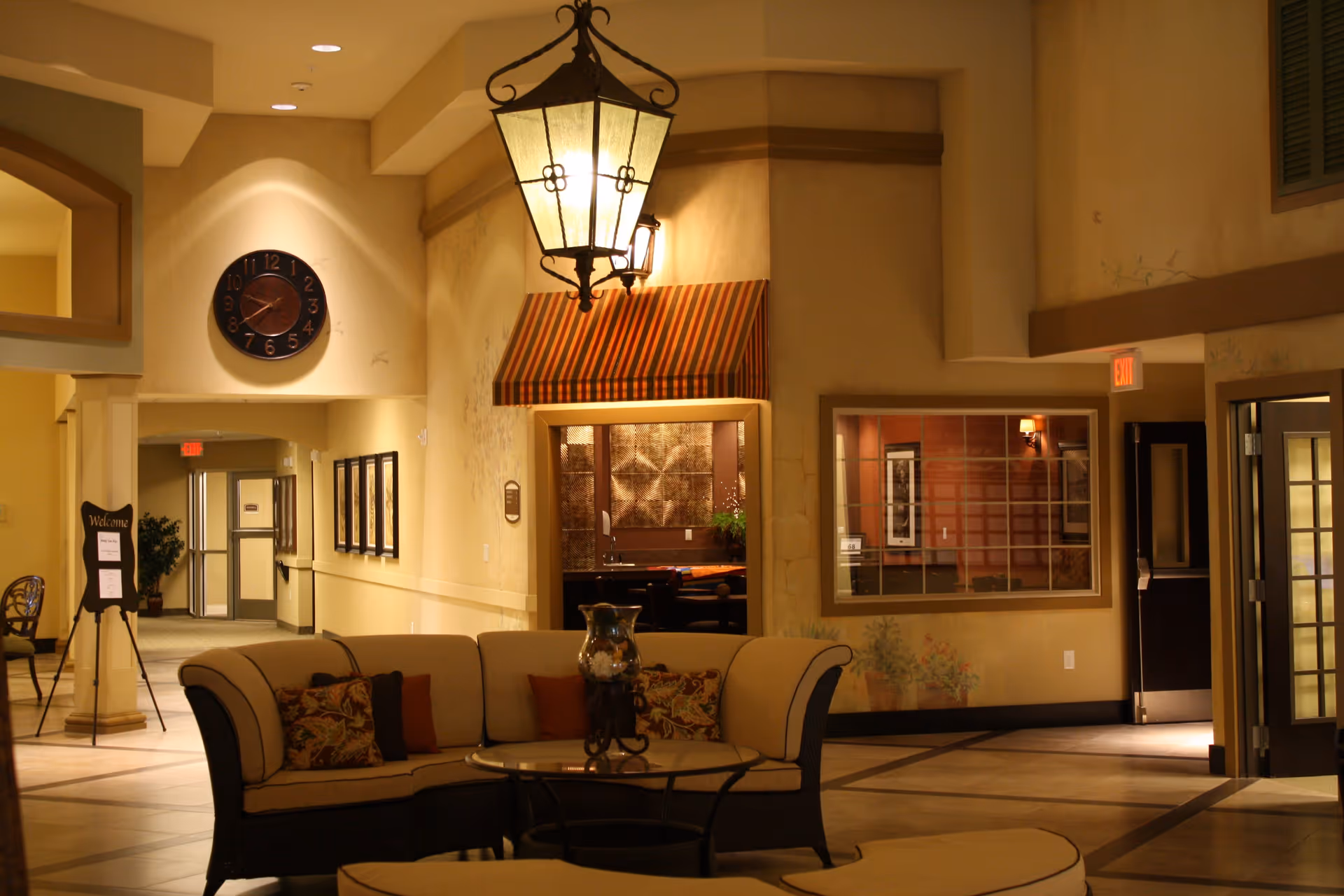 Interior view of a senior living facility lounge area with a curved beige sofa, decorative pillows, a glass-top coffee table, and a large hanging lantern light fixture. The walls are painted in warm tones with a striped awning above a doorway and a clock mounted on the wall. There is a hallway leading to other rooms and an exit sign above a door.
