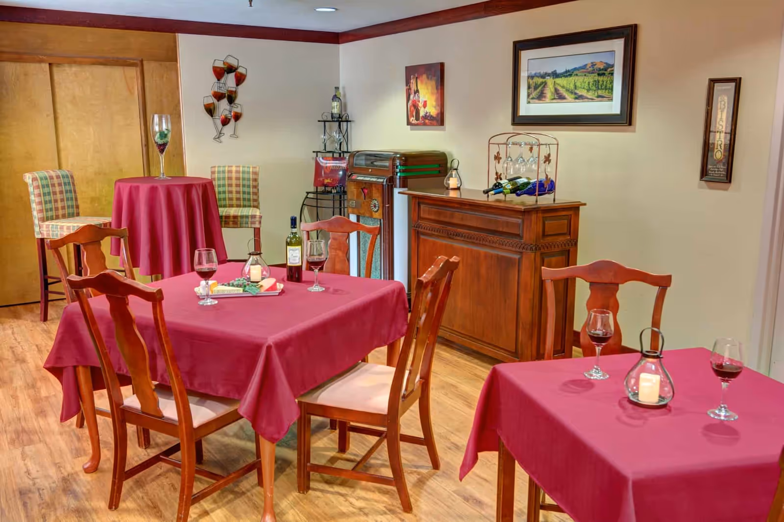 Small dining/lounge area with tables draped in red tablecloths, wooden chairs, wine glasses, and a wooden sideboard with decor against the wall.