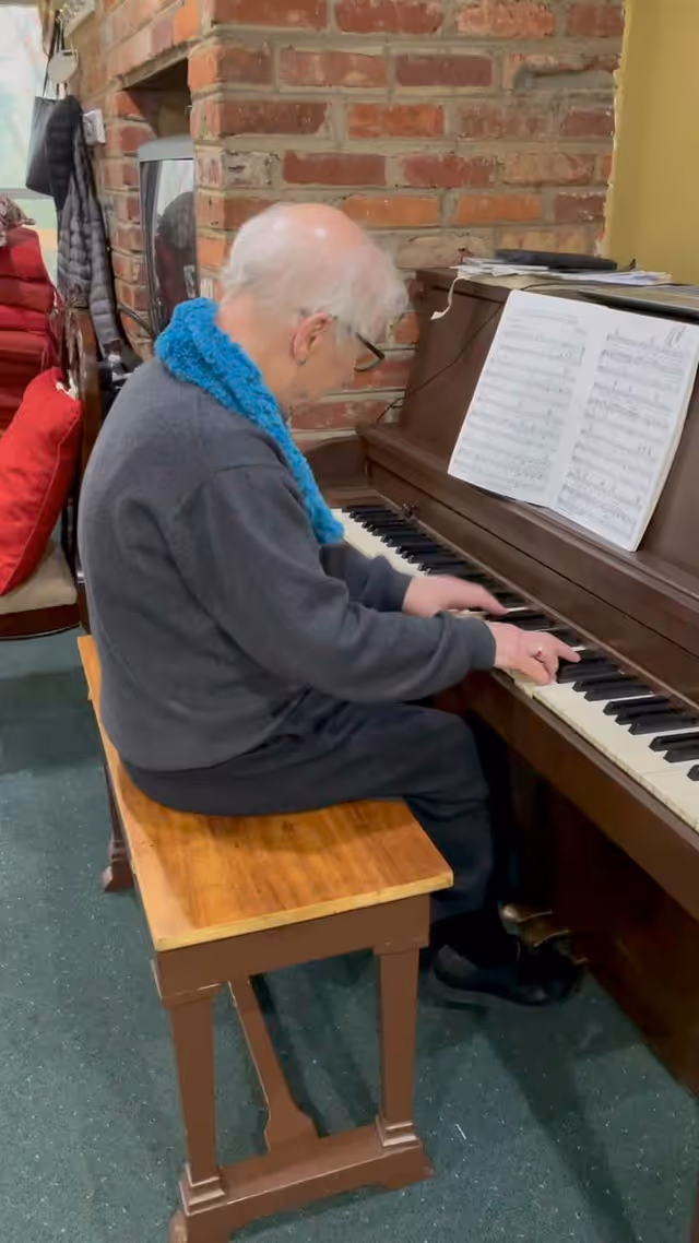 An elderly person with white hair and glasses is seated on a wooden bench playing a brown upright piano. Sheet music is placed on the piano stand. The background features a brick wall and some red cushions on a chair.
