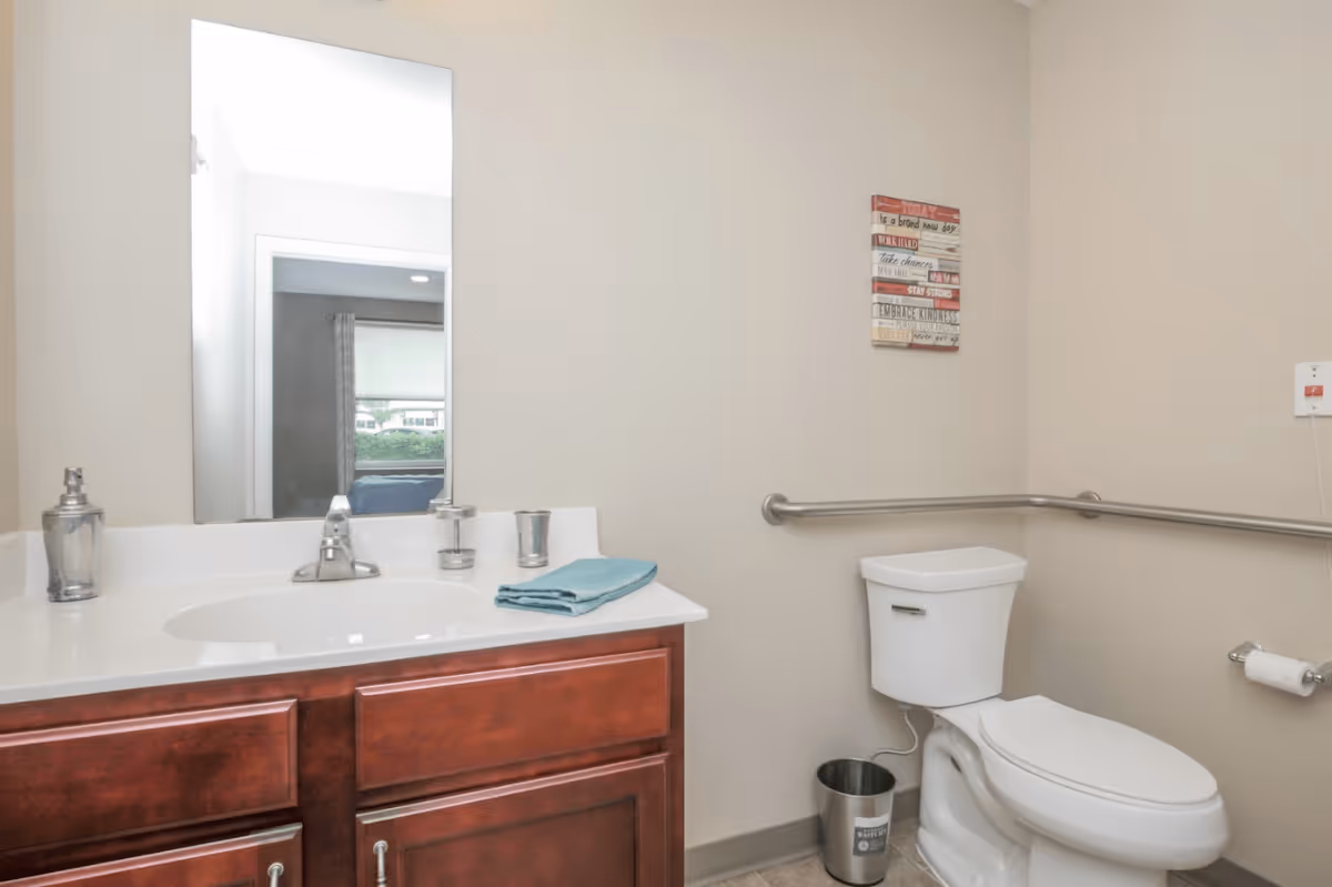 A clean bathroom with a white toilet, a silver grab bar on the wall, a small metal trash can, and a wooden vanity with a white countertop. On the countertop are a silver soap dispenser, a folded teal towel, and a small silver container. A rectangular mirror hangs above the sink, reflecting part of a bedroom with a window and curtains. A decorative sign with motivational phrases is mounted on the wall above the toilet.