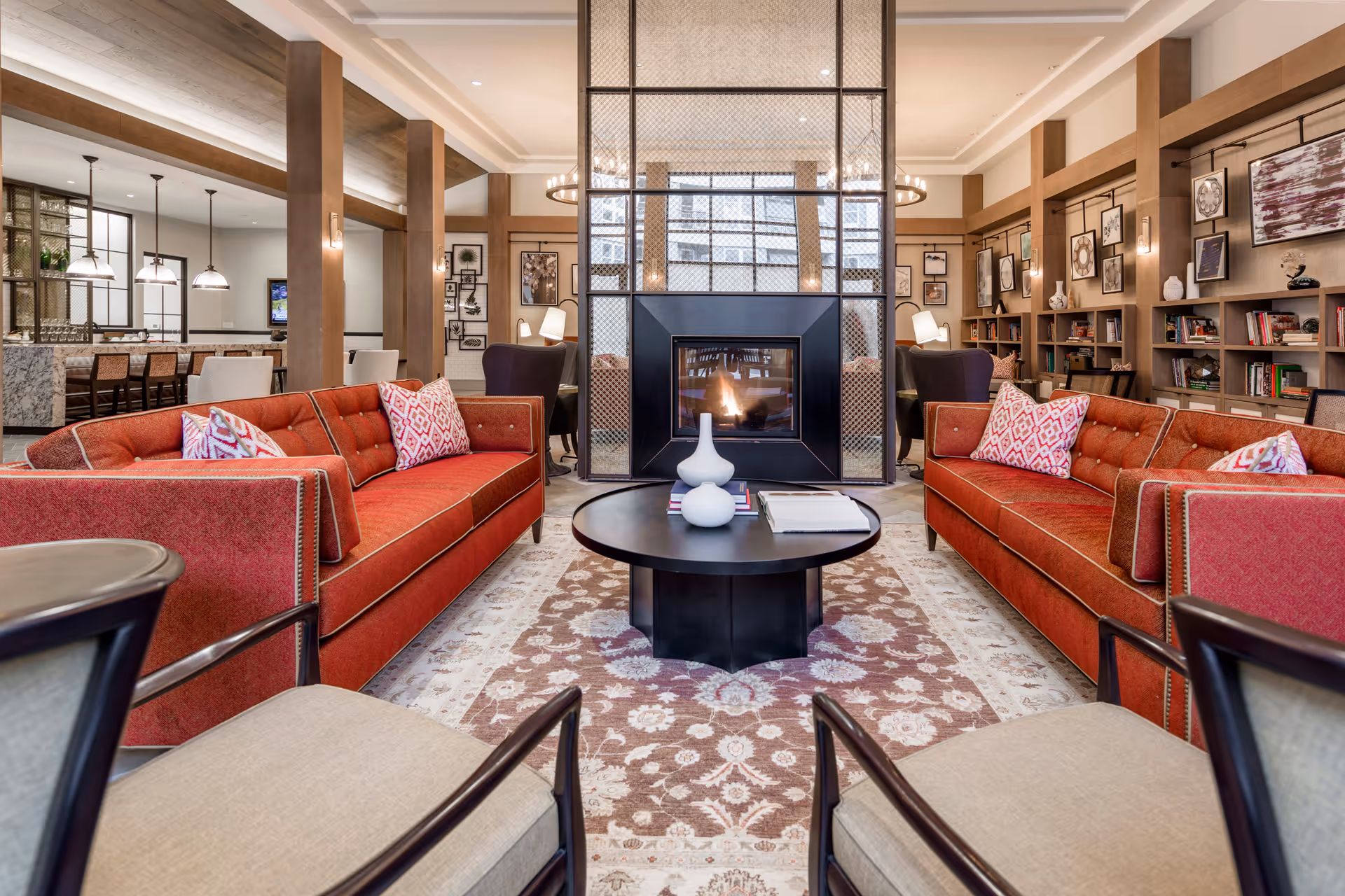 A cozy and stylish living room area featuring two red sofas with patterned pillows facing each other across a round black coffee table. The room has a central modern fireplace with a glass and metal screen, flanked by bookshelves filled with books and decorative items. The space is well-lit with ceiling lights and has wooden beams and columns, creating a warm and inviting atmosphere.