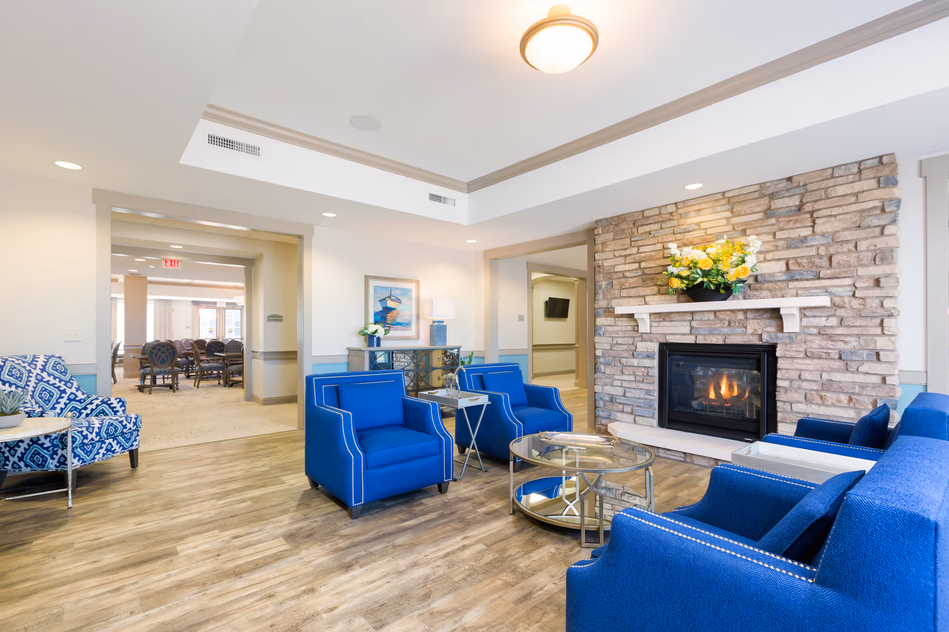 A bright and inviting senior living facility common area featuring a stone fireplace with a floral arrangement on the mantel. The room has blue upholstered chairs and a sofa arranged around a glass coffee table on wood flooring. In the background, there is a hallway leading to another room with dining tables and chairs.