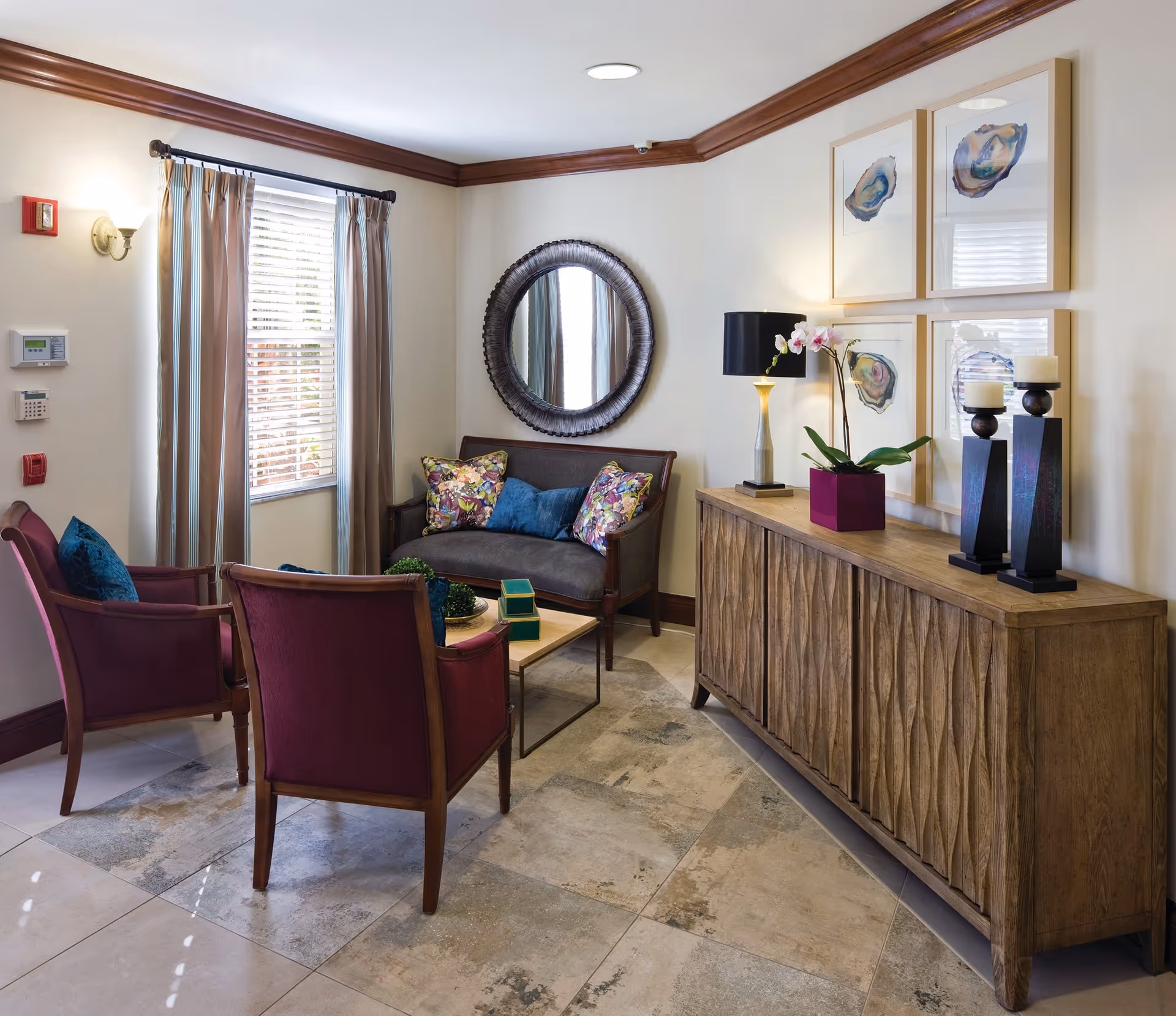 A cozy sitting area with two purple armchairs and a dark gray loveseat adorned with colorful pillows. A wooden coffee table with decorative items sits in the center on a tiled floor. A wooden sideboard with a textured front holds a lamp, a purple potted orchid, and two candle holders. Four framed abstract art pieces hang above the sideboard. A round mirror is mounted on the wall behind the loveseat, and a window with beige curtains lets in natural light.