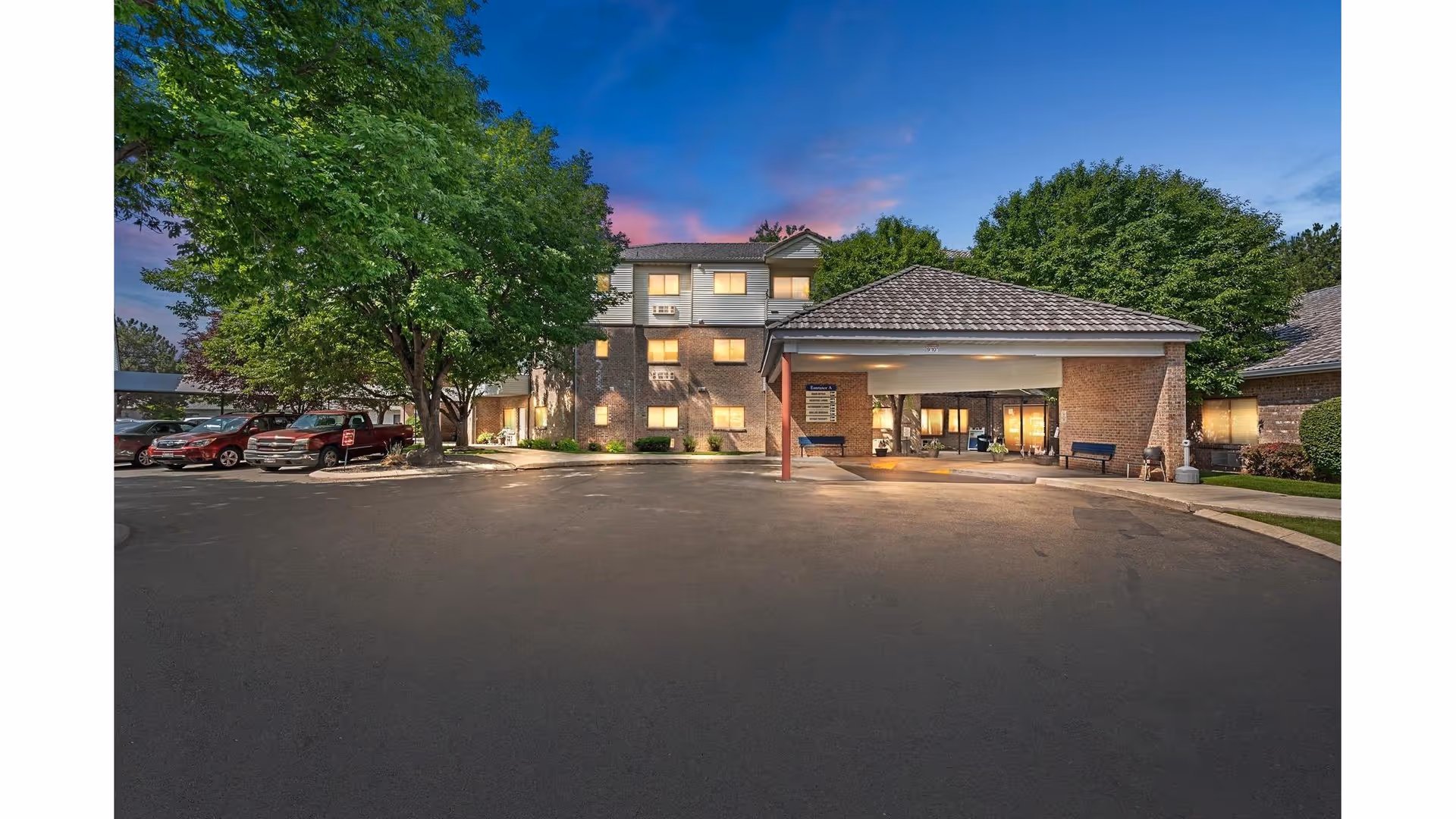 Exterior view of Karcher Senior Living facility at dusk, showing a three-story building with illuminated windows, a covered entrance with benches, a parking area with several cars, and large green trees surrounding the property.