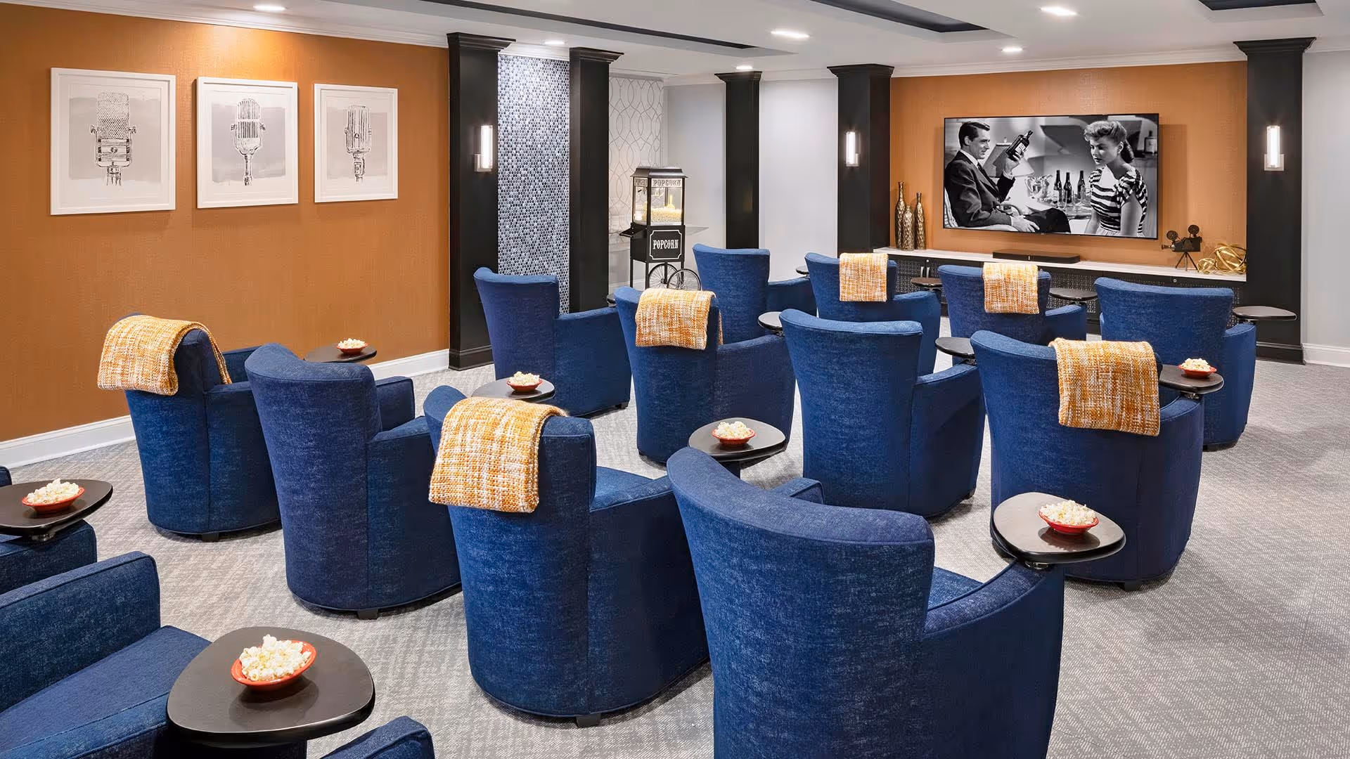 Media room with rows of blue armchairs facing a wall-mounted TV, each chair with an orange throw and a small side table holding a bowl of popcorn.