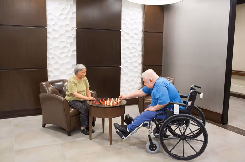 Two elderly individuals playing chess in a comfortable indoor seating area. One person is seated in a brown armchair, and the other is in a wheelchair. The background features a modern wall design with dark wood panels and textured white sections.
