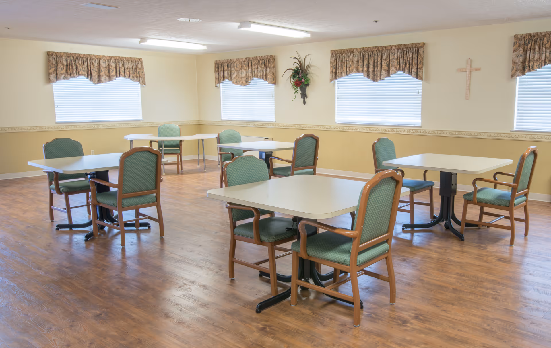 A bright communal dining room with several tables and green-upholstered chairs on a wood floor.